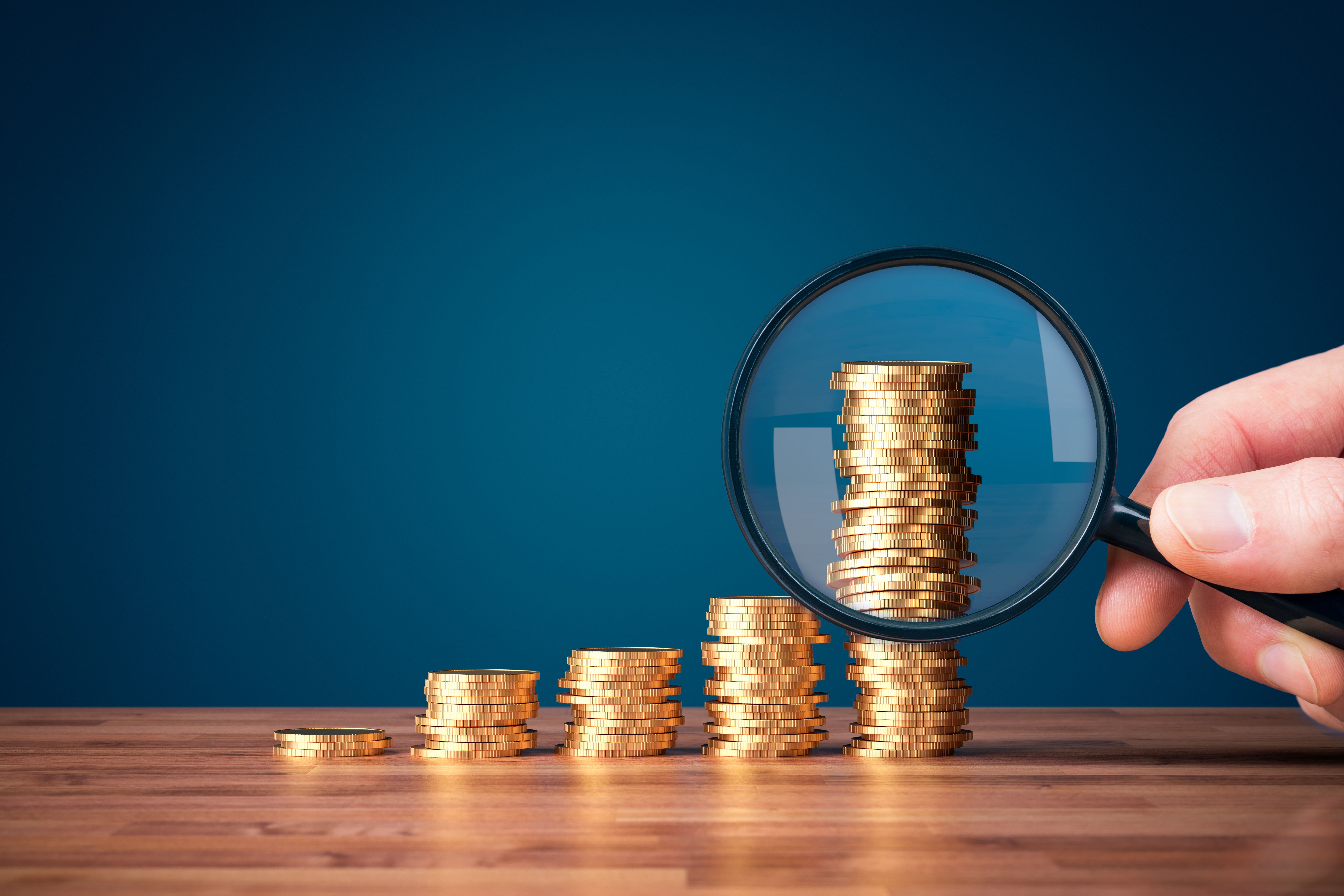 Different size piles of coin being investigated with a magnifying glass | Image Credit: jirsak - stock.adobe.com