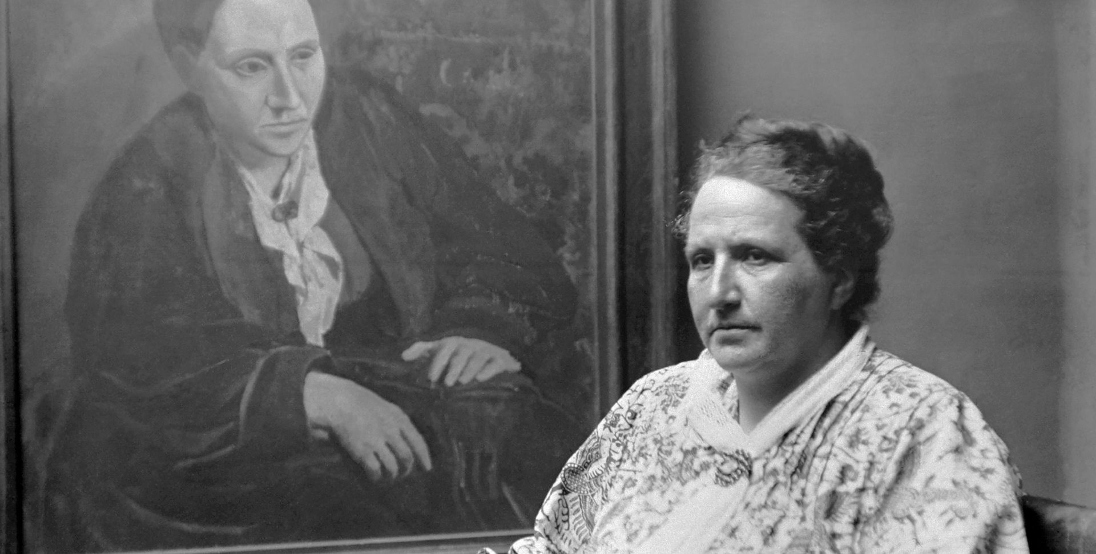 Black and white photograph of a woman wearing a white floral pattern outfit on the right with a potrait in the background done by Picasso of her. 