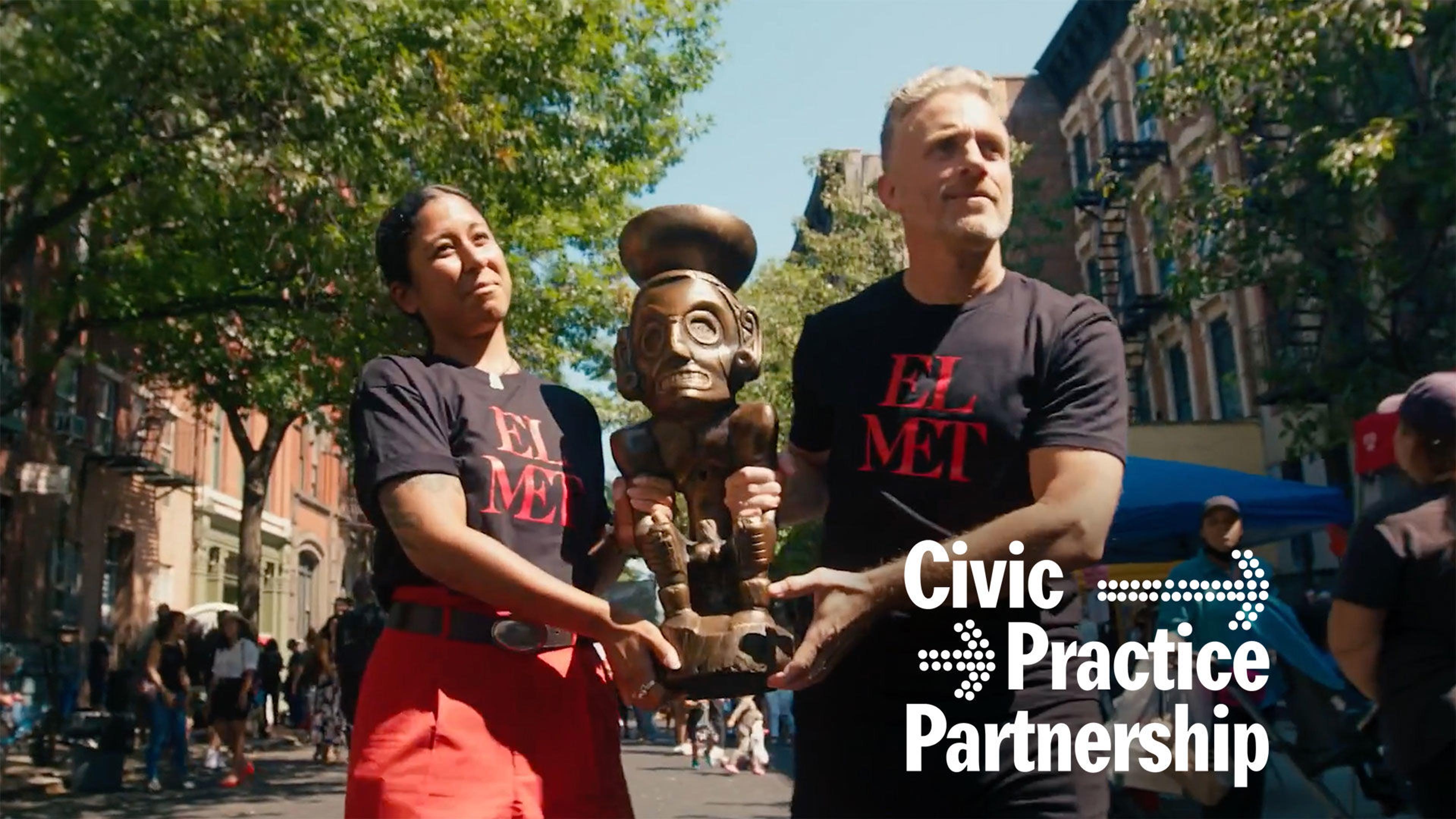 Artist Miguel Luciano and a woman wearing red pants carrying the Zemí Cohoba stand through the streets of El Barrio