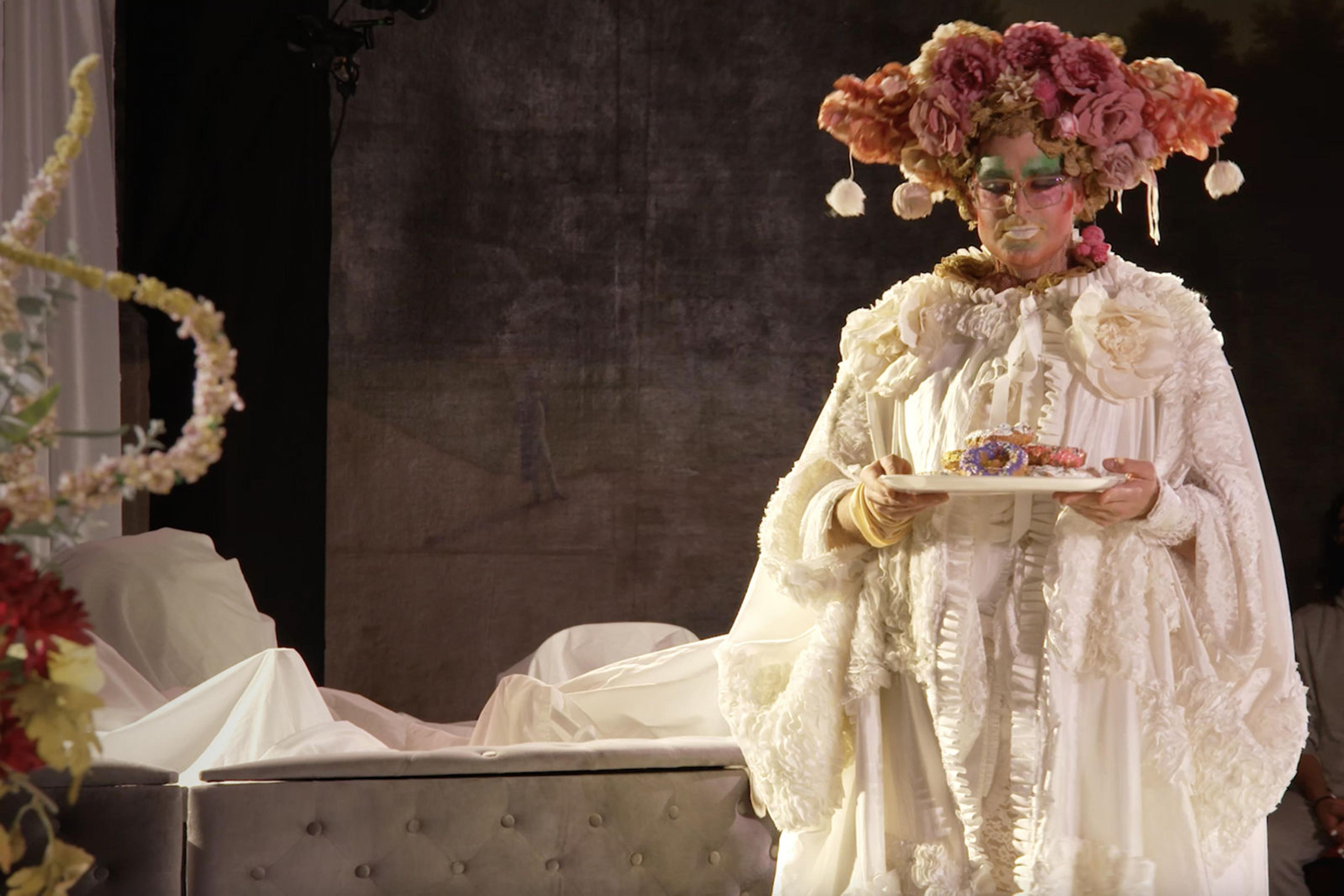 A figure in bold, colorful makeup and elaborate costuming stands holding a platter with donuts. Their white dress is ruffled, and they're wearing a headdress made of pink flowers.