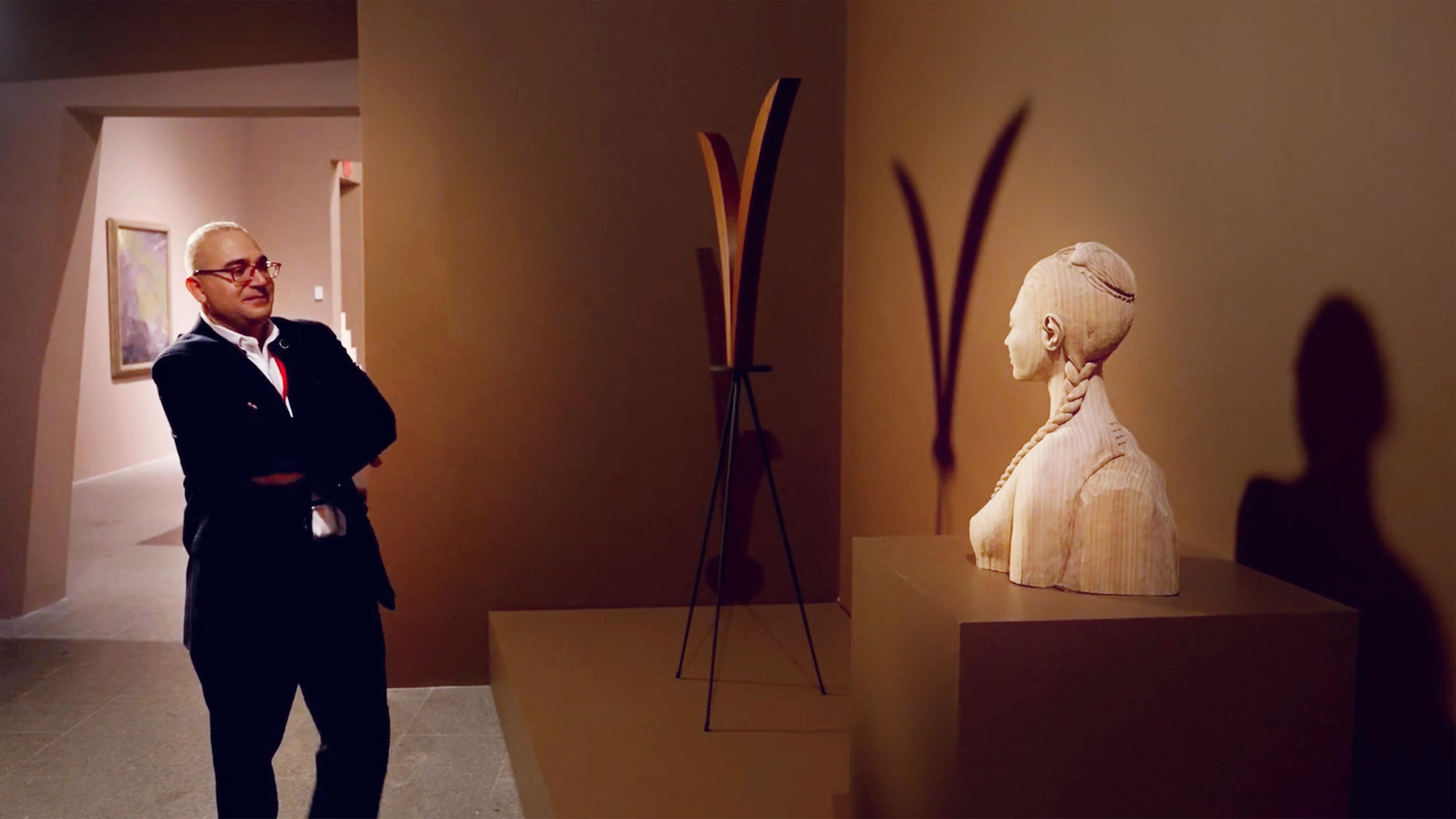Met security guard Armia Khalil crosses his arms and smiles while standing across from his artwork in gallery, a carved bust of a woman with a scarab in her hair.